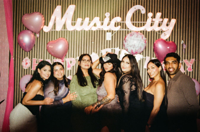Birthday party photo of friends posing under an illuminated "Music City" sign with pink heart balloons and a birthday banner