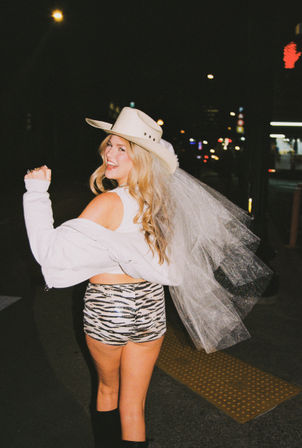 Party-ready woman in a cowboy hat and sparkly bridal veil, zebra-print shorts and off-shoulder jacket on a neon-lit city street at night.