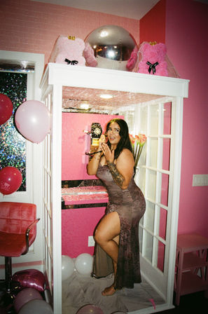 Person posing in a pink-themed decorative phone booth holding a retro rotary phone, surrounded by balloons, plush pink decor, glitter tiles and flowers — festive indoor party scene