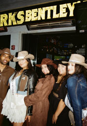 Five friends in cowboy hats pose for a night-out photo under a lit marquee outside a bar, one wearing a birthday sash.