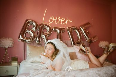 Playful bride-to-be in a veil and sequin dress lounging on a pink bed, sipping a drink beneath a neon "Lover" sign and metallic "BRIDE" balloons