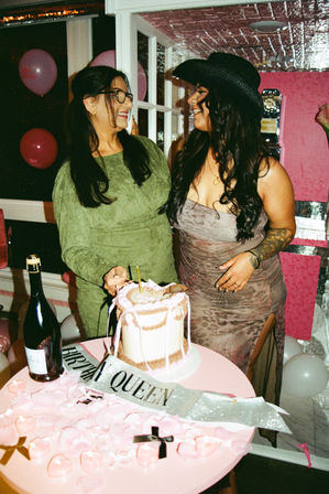 Two women laughing at an indoor birthday party beside a pink frosted cake and a sparkly "BIRTHDAY QUEEN" sash, with pink balloons, rose petals and a champagne bottle; one wears a black cowboy hat and patterned dress, the other a green dress and glasses.