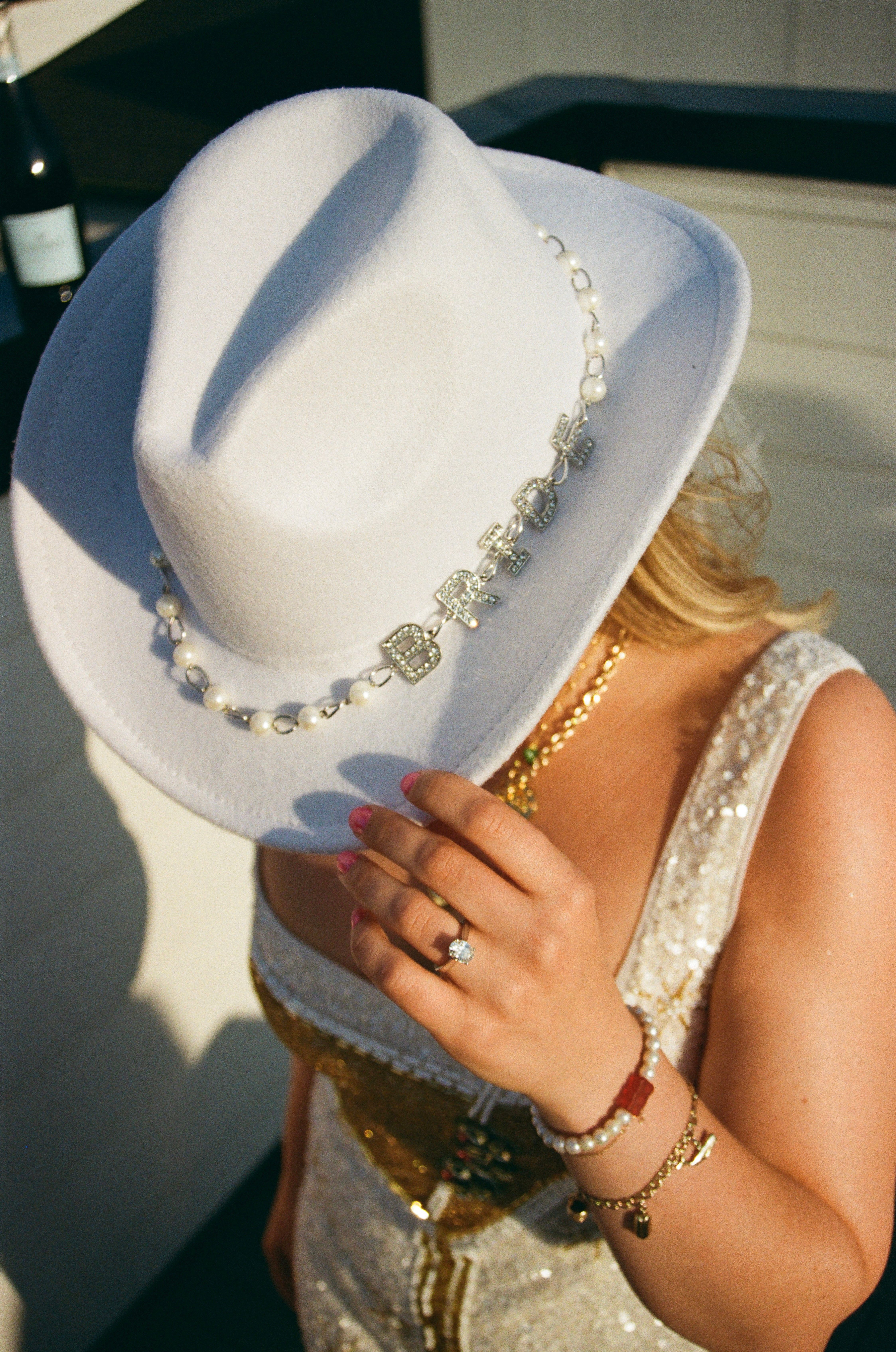 Sunlit white felt cowboy hat with pearl and rhinestone 'BRIDE' chain, hand adjusting brim to show engagement ring, gold jewelry and sequined bridal dress.