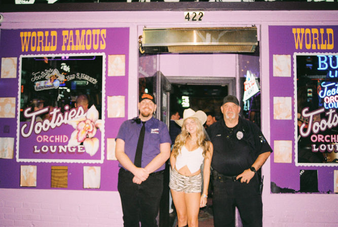 Three smiling people — a young woman in a cowboy hat and shorts between two uniformed staff — pose outside a purple neon-lit bar entrance marked 422.