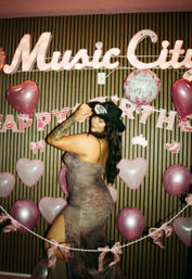 Woman in lace evening dress and black hat posing at a birthday photo booth with pink heart balloons and a neon "Music City" sign