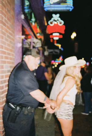 Officer handcuffing a woman in a cowboy hat and veil on a neon-lit downtown nightlife street at night