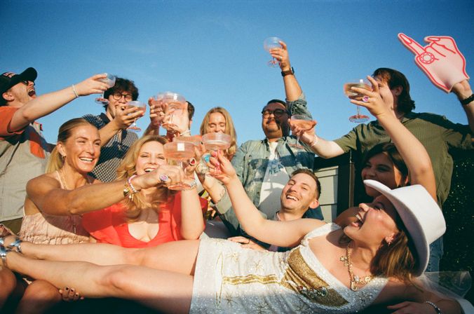 Friends at an outdoor sunny party cheering and clinking champagne coupes while lifting a woman in a white dress and cowboy hat in a playful toast.