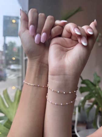Close-up of two manicured hands and wrists with glossy pink and French-tip nails wearing delicate gold beaded chain bracelets, soft natural light with a blurred window and green plants in the background.