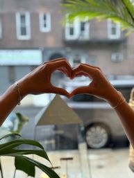 Two hands forming a heart shape against a window overlooking an urban street with a brick building and parked car, framed by leafy plant leaves and delicate bracelets on the wrists.