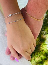 Close-up of two hands holding outdoors on green grass, featuring delicate silver bracelets — one with tiny blue beads and a small round charm — and a gold chain bracelet; one hand sports bright purple nail polish.