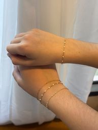 Close-up of two wrists wearing delicate gold chain bracelets with tiny white beads, dainty jewelry styled against a soft white curtain background