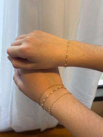 Close-up of two wrists wearing delicate gold chain bracelets with tiny white beads, dainty jewelry styled against a soft white curtain background