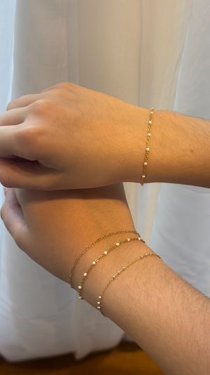 Close-up of two wrists wearing delicate gold chain bracelets with tiny white beads, dainty jewelry styled against a soft white curtain background