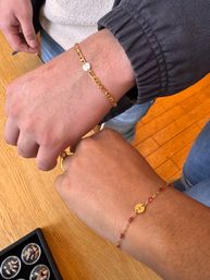 Close-up of two wrists on a wooden tabletop wearing matching delicate gold chain bracelets with small round charms—one bracelet has small red beads—photo taken near a jewelry display.