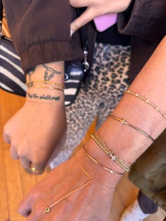 Close-up of two wrists wearing stacked gold bracelets and a delicate hand chain; left wrist shows a small longhorn tattoo and script, zebra-striped bag and leopard-print pants visible in the background.
