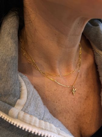 Close-up of a neck wearing layered gold necklaces, one with a small angel pendant, over a gray zip-up hoodie.