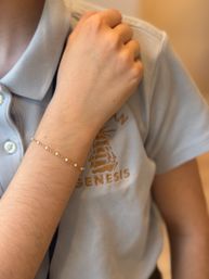Close-up of a wrist wearing a delicate gold chain bracelet dotted with tiny pearl beads, hand resting on a light-blue polo shirt with embroidered logo.