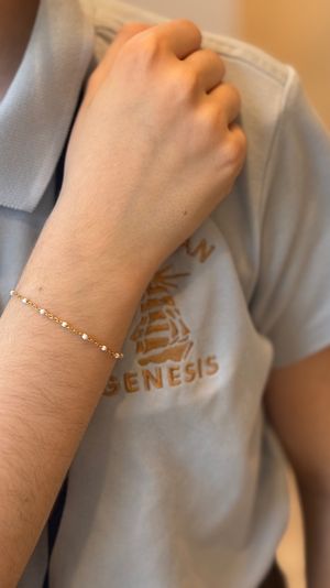 Close-up of a wrist wearing a delicate gold chain bracelet dotted with tiny pearl beads, hand resting on a light-blue polo shirt with embroidered logo.