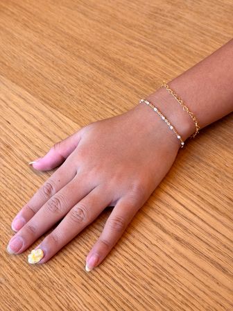 Close-up of a hand resting on a wooden table wearing two delicate gold chain bracelets and yellow 3D flower nail art on the thumb.