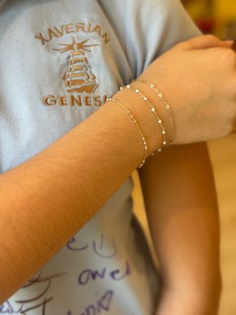 Close-up of a forearm wearing three delicate gold chain bracelets with tiny white beads over a light-blue embroidered shirt with a stitched crest