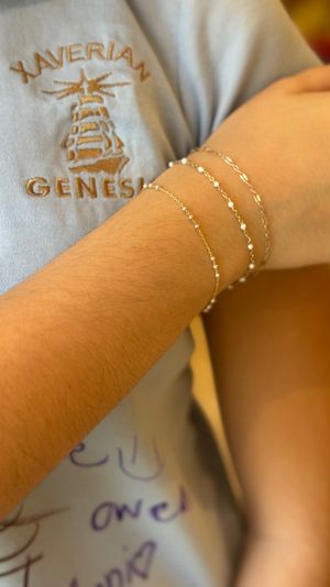 Close-up of a forearm wearing three delicate gold chain bracelets with tiny white beads over a light-blue embroidered shirt with a stitched crest