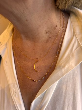 Close-up of a neckline wearing layered gold necklaces — a delicate crescent moon pendant and beaded gold chain over a white button-down shirt, warm light, fashion jewelry detail.