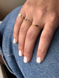 Close-up of a hand resting on blue denim wearing three dainty gold chain rings — a small disc, a tiny heart, and a pink-red rectangular stone — with short white manicure featuring subtle floral and polka-dot nail art.