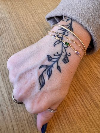 Close-up of a hand on a wooden table showing a black floral vine wrist tattoo, a gold bangle and two delicate gold chain bracelets with green and purple rectangular stones, plus stacked rings.