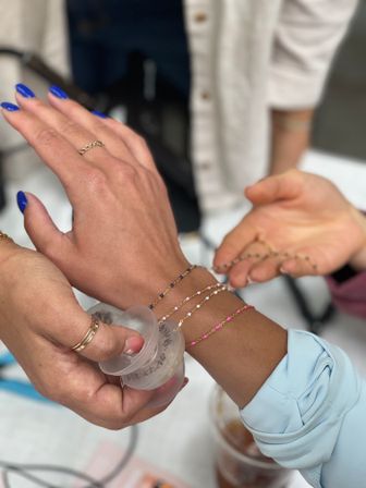 Close-up of hands trying on delicate beaded bracelets — three stacked pink, gold and navy bead chains on a wrist, bright blue nail polish and gold rings, jewelry fitting scene.