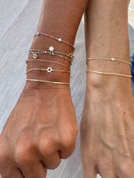 Close-up of two tanned wrists with stacked delicate gold chains and colorful beaded bracelets featuring tiny sun and star-shaped charms on a light linen background — stylish summer wrist jewelry.