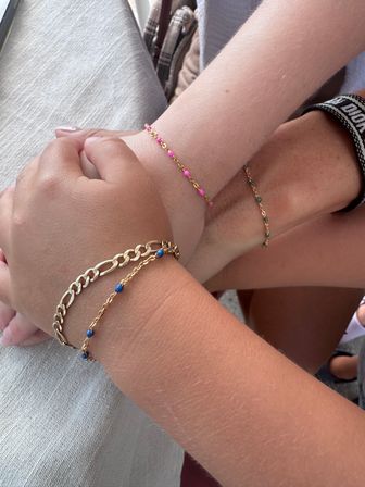 Three joined wrists wearing stacked delicate gold chain bracelets with tiny pink, blue, and green beads in a casual setting