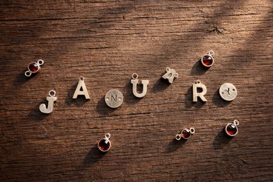 Metal letter charms arranged to spell "JANUARY" on a rustic wooden table, scattered with small red gemstone pendant charms.