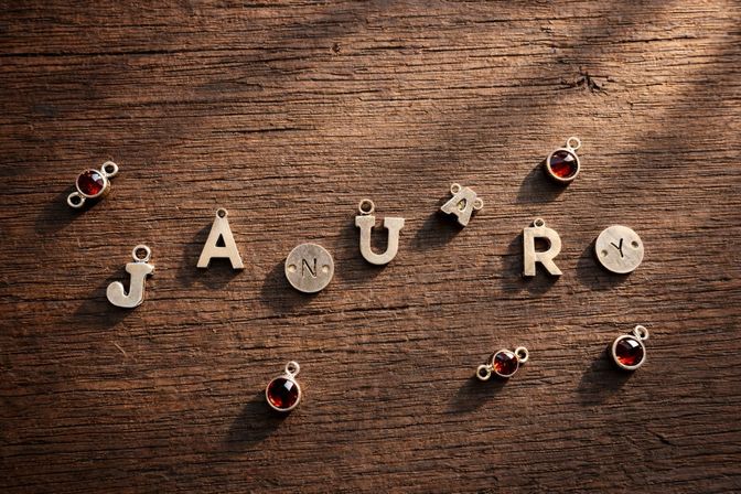 Metal letter charms arranged to spell "JANUARY" on a rustic wooden table, scattered with small red gemstone pendant charms.