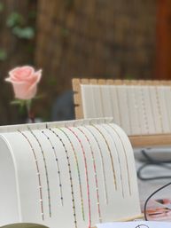 Delicate beaded chain bracelets in gold, silver and bright neon tones draped on a white curved display stand, with a soft-focus pink rose and wooden jewelry tray in an outdoor boutique market setting.