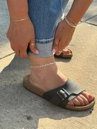 Close-up of cuffed blue jeans and hands adjusting the hem to show layered silver anklets and coral-painted toes in a black leather slide sandal with cork sole on sunlit concrete, wearing multiple gold and silver rings and bracelets.