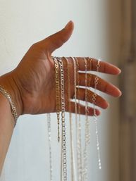 Close-up of a hand holding assorted gold and silver chain necklaces with varied link styles, draping against a neutral indoor wall — minimalist jewelry display