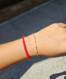 Sunlit wrist wearing a braided red string bracelet and a delicate gold chain, photographed over concrete pavement