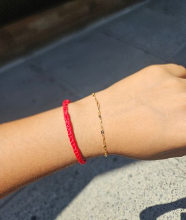 Sunlit wrist wearing a braided red string bracelet and a delicate gold chain, photographed over concrete pavement