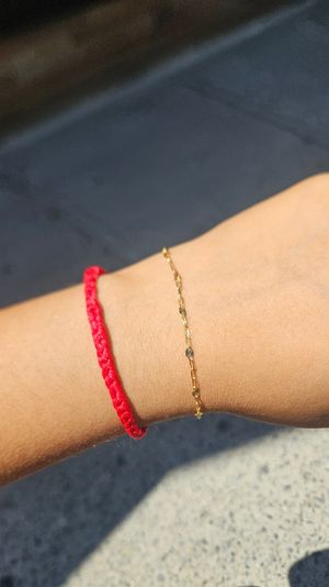Sunlit wrist wearing a braided red string bracelet and a delicate gold chain, photographed over concrete pavement