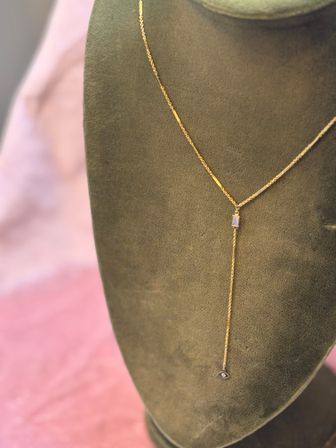Delicate gold lariat necklace with a small rectangular opalescent bead and tiny eye charm on an olive-green velvet bust, soft pink backdrop.