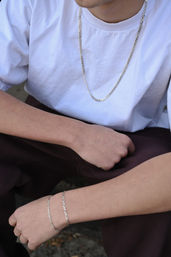 Close-up of a person in a white T-shirt wearing a silver chain necklace and two matching silver bracelets, arms resting on dark pants for a minimalist casual streetwear look