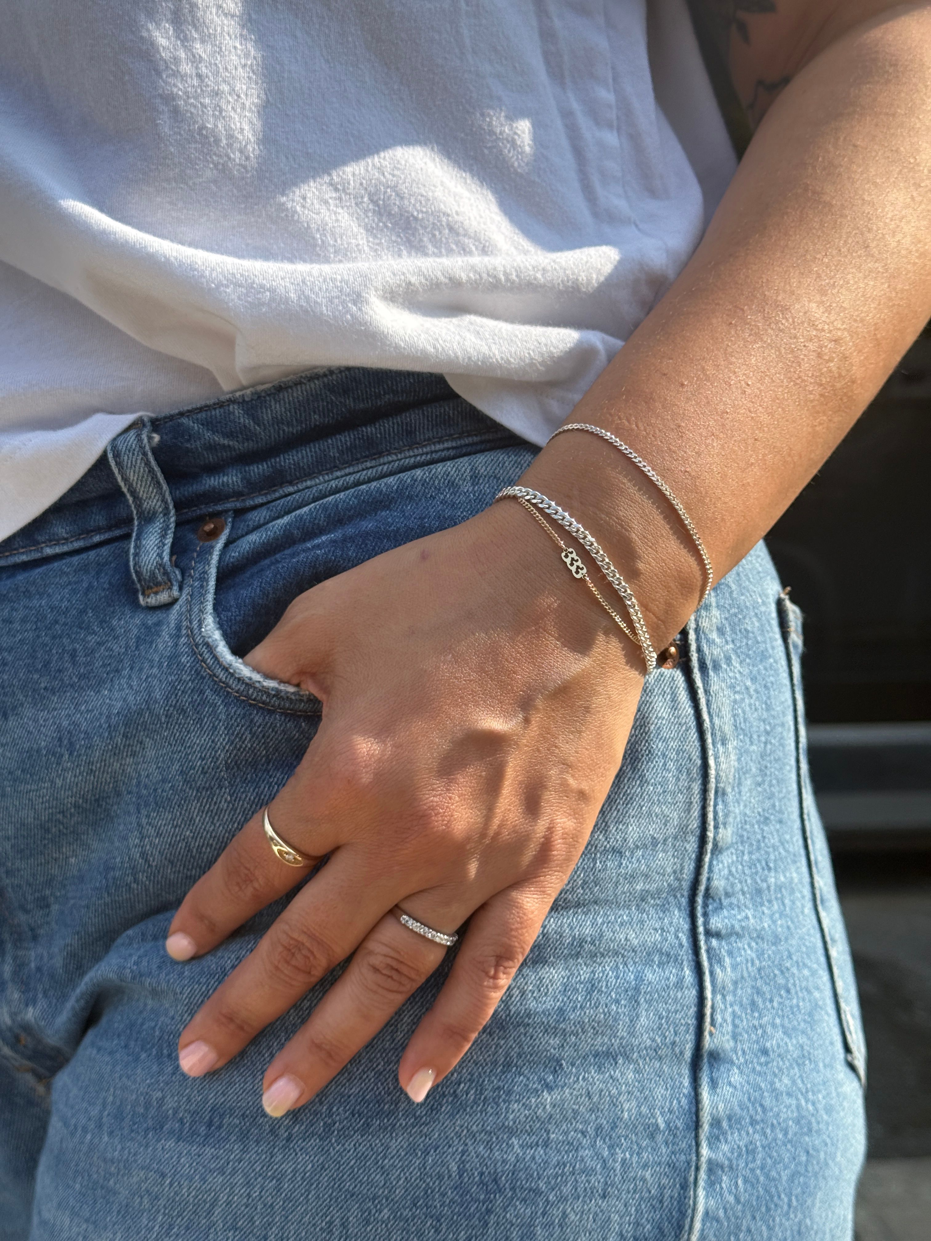 Close-up of hand tucked in blue denim pocket wearing delicate silver chain bracelets and two rings, casual white t-shirt — minimalist summer street-style jewelry shot.