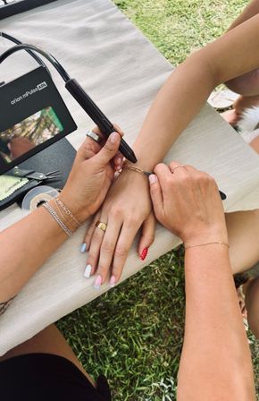 Close-up of two people's hands outdoors as one person uses a handheld tool to fasten a delicate gold chain bracelet on the other's wrist; manicured nails with pastel and red polka-dot polish, rings and bracelets, small pliers and tape on a tablecloth over grass.