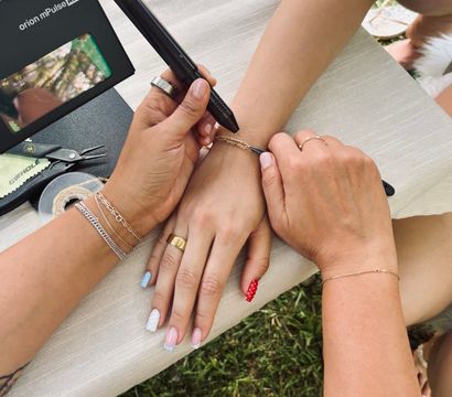 Close-up of two people's hands outdoors as one person uses a handheld tool to fasten a delicate gold chain bracelet on the other's wrist; manicured nails with pastel and red polka-dot polish, rings and bracelets, small pliers and tape on a tablecloth over grass.