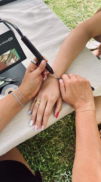 Close-up of two people's hands outdoors as one person uses a handheld tool to fasten a delicate gold chain bracelet on the other's wrist; manicured nails with pastel and red polka-dot polish, rings and bracelets, small pliers and tape on a tablecloth over grass.