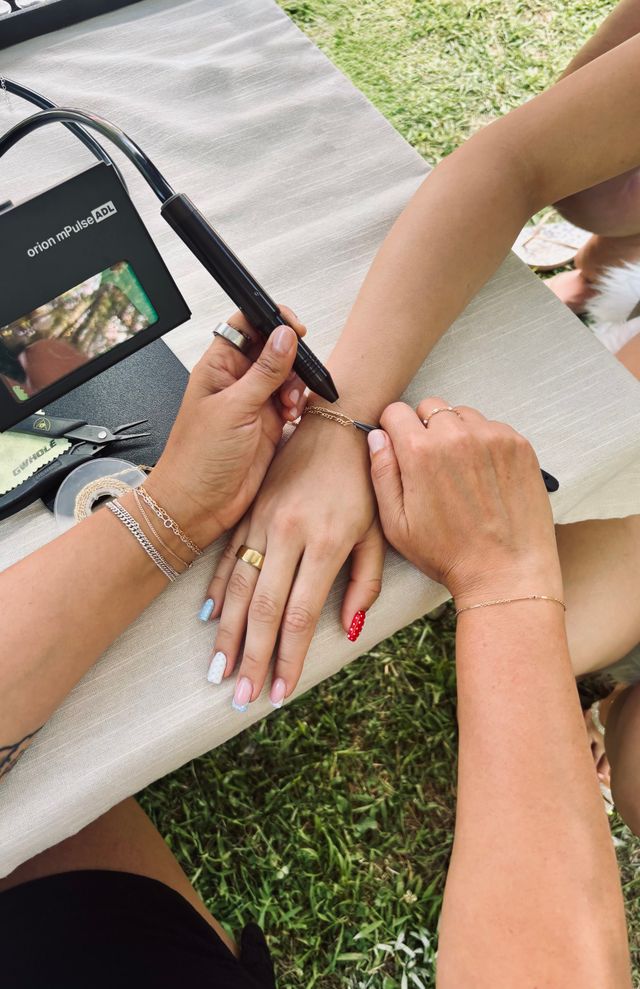 Close-up of two people's hands outdoors as one person uses a handheld tool to fasten a delicate gold chain bracelet on the other's wrist; manicured nails with pastel and red polka-dot polish, rings and bracelets, small pliers and tape on a tablecloth over grass.
