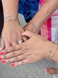 Three hands stacked outdoors showcasing bright coral nail polish, a sparkling diamond ring, and delicate gold and blue beaded bracelets on summery pavement.