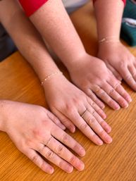 Close-up of three hands lined side-by-side on a wooden table, each wearing thin rings and delicate bracelets — minimalist jewelry styling