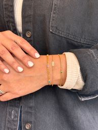 Close-up of a wrist wearing three delicate gold chain bracelets with small colorful gemstones and a tiny charm, stacked over a cream sweater cuff and dark denim jacket, with neatly manicured white nails — casual jewelry detail shot.
