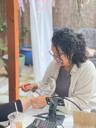 Cozy backyard pedicure scene: a nail technician with curly hair and glasses tending to a client’s foot near a sneaker at a patio nail station with tools, portable lamp, iced coffee and potted plants.